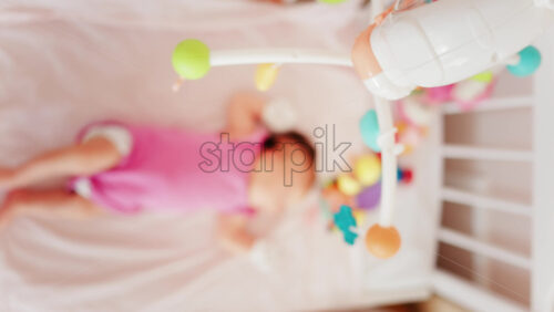 Baby lying on a bed in a pink outfit, looking up at a colourful hanging mobile toy - Starpik Stock