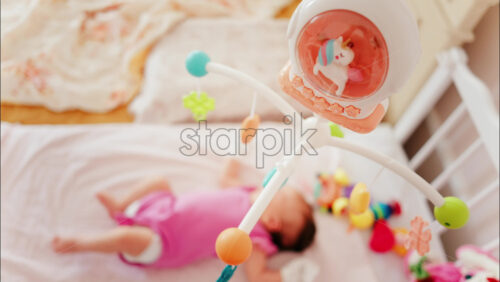 Baby lying on a bed in a pink outfit, looking up at a colourful hanging mobile toy - Starpik Stock