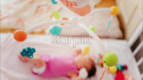 Baby lying on a bed in a pink outfit, looking up at a colourful hanging mobile toy - Starpik Stock