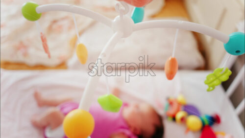 Baby lying on a bed in a pink outfit, looking up at a colourful hanging mobile toy - Starpik Stock