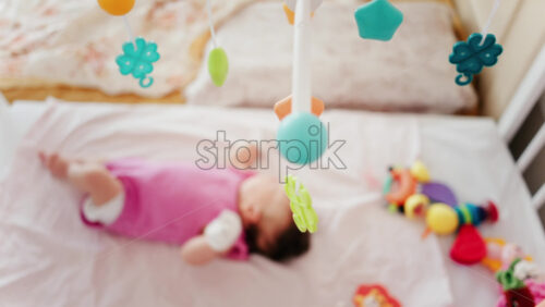 Baby lying on a bed in a pink outfit, looking up at a colourful hanging mobile toy - Starpik Stock