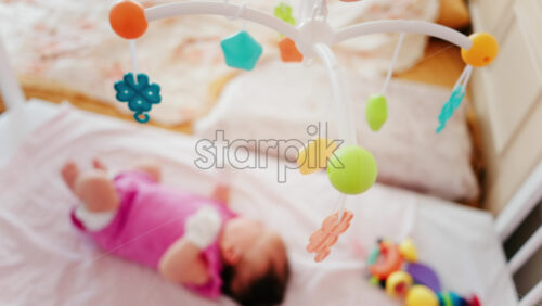 Baby lying on a bed in a pink outfit, looking up at a colourful hanging mobile toy - Starpik Stock