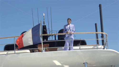 Cannes, France – July 10, 2025: Woman in a blue suit leaning on the aft rail of a yacht under clear blue sky with the French flag waving at the stern - Starpik Stock