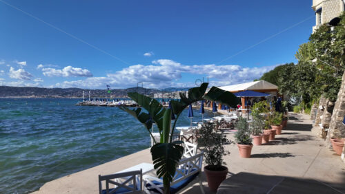 Cannes, France – July 12, 2025: A peaceful seaside walkway lined with terracotta pots, banana leaves and umbrellas overlooking blue water - Starpik Stock