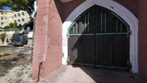 Cannes, France – July 12, 2025: Visitors stroll a narrow seawall beside a grand waterfront hotel and beach club with turquoise water laps against the rocks - Starpik Stock