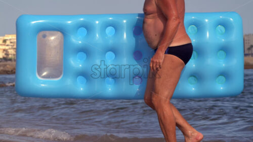 Cannes, France – July 9, 2025: Close up of an adult man in swim briefs carrying a bright blue air mattress along the shoreline - Starpik Stock