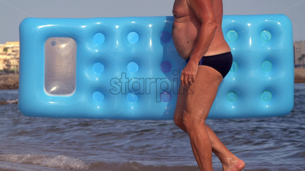 Cannes, France – July 9, 2025: Close up of an adult man in swim briefs carrying a bright blue air mattress along the shoreline - Starpik Stock
