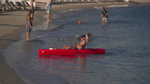 Cannes, France – June 22, 2025: Man kayaking in a bright red kayak near the shore, with few people playing with a ball on the sand, while a dog frolics happily close to the water’s edge - Starpik Stock
