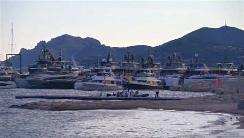 Cannes, France – June 22, 2025: People walking near boats docked in the Cannes harbour with mountains in the background in the evening - Starpik Stock