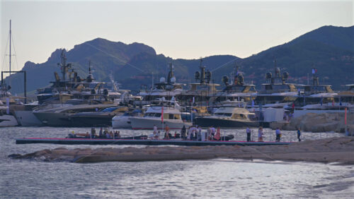 Cannes, France – June 22, 2025: People walking near boats docked in the Cannes harbour with mountains in the background in the evening - Starpik Stock