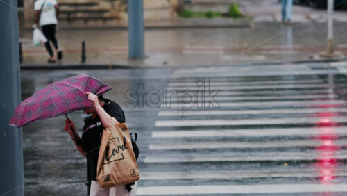 Chisinau, Moldova – June 11, 2025: Woman crossing the street while holding an umbrella in the wind and rain - Starpik Stock