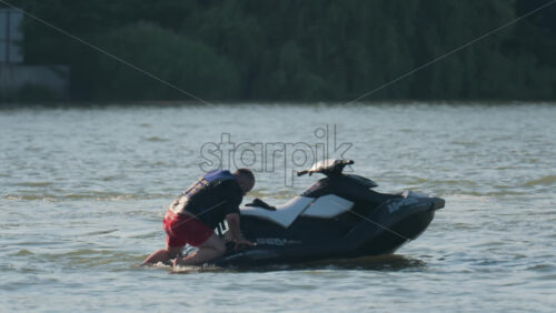 Chisinau, Moldova – June 17, 2025: A man in a black life vest climbing a black jet ski from a calm body of water - Starpik Stock