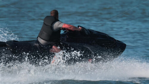 Chisinau, Moldova – June 17, 2025: A man in a black life vest riding a black jet ski at high speed across a calm body of water - Starpik Stock