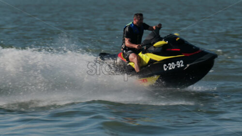Chisinau, Moldova – June 17, 2025: A man in a black life vest riding a black jet ski at high speed across a calm body of water - Starpik Stock
