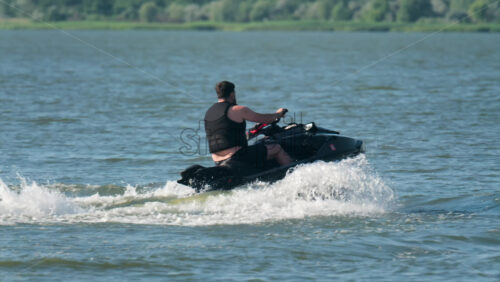 Chisinau, Moldova – June 17, 2025: A man in a black life vest riding a black jet ski at high speed across a calm body of water - Starpik Stock