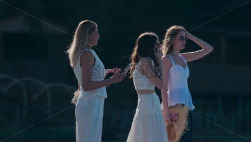 Chisinau, Moldova – June 17, 2025: Three fashionable women in white clothes standing outdoors, talking and holding mobile phones - Starpik Stock