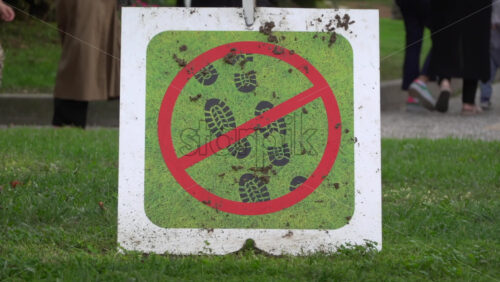 Close-up of a No Walking on Grass sign placed on a lawn with pedestrians walking in the background - Starpik Stock