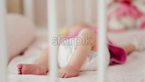 Close up of a baby’s legs and diaper as the infant rests in a crib, seen through the crib bars - Starpik Stock