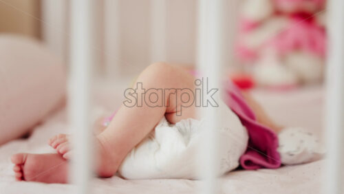 Close up of a baby’s legs and diaper as the infant rests in a crib, seen through the crib bars - Starpik Stock