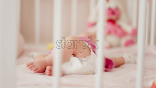 Close up of a baby’s legs and diaper as the infant rests in a crib, seen through the crib bars - Starpik Stock