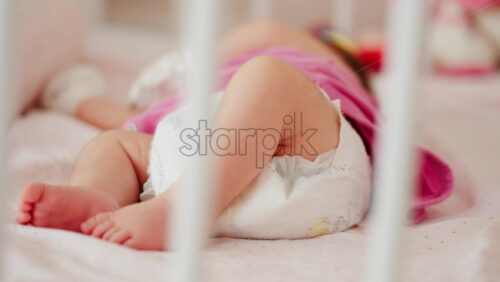 Close up of a baby’s legs and diaper as the infant rests in a crib, seen through the crib bars - Starpik Stock