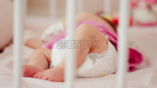 Close up of a baby’s legs and diaper as the infant rests in a crib, seen through the crib bars - Starpik Stock