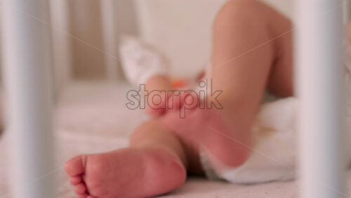 Close up of a baby’s legs and diaper as the infant rests in a crib, seen through the crib bars - Starpik Stock