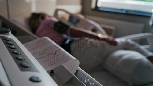 Close-up of a fetal monitor in a doctor’s office, displaying fetal heart rate and uterine contraction data during a prenatal check-up with a blurred view of a pregnant woman lying in a hospital bed - Starpik Stock