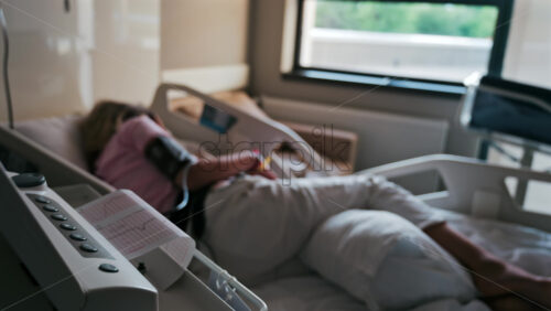 Close-up of a fetal monitor in a doctor’s office, displaying fetal heart rate and uterine contraction data during a prenatal check-up with a blurred view of a pregnant woman lying in a hospital bed - Starpik Stock
