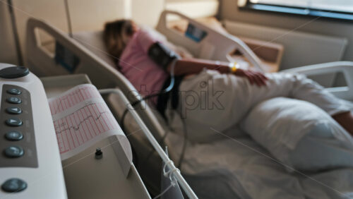 Close-up of a fetal monitor in a doctor’s office, displaying fetal heart rate and uterine contraction data during a prenatal check-up with a blurred view of a pregnant woman lying in a hospital bed - Starpik Stock