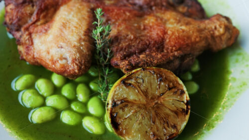 Close-up of a gourmet roasted chicken thigh served with edamame beans, a drizzle of herb-infused green sauce, and a charred lime half - Starpik Stock