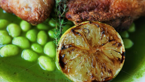 Close-up of a gourmet roasted chicken thigh served with edamame beans, a drizzle of herb-infused green sauce, and a charred lime half - Starpik Stock