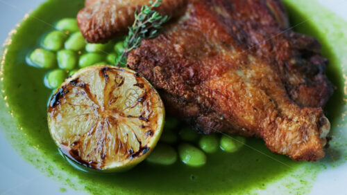 Close-up of a gourmet roasted chicken thigh served with edamame beans, a drizzle of herb-infused green sauce, and a charred lime half - Starpik Stock