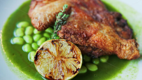 Close-up of a gourmet roasted chicken thigh served with edamame beans, a drizzle of herb-infused green sauce, and a charred lime half - Starpik Stock