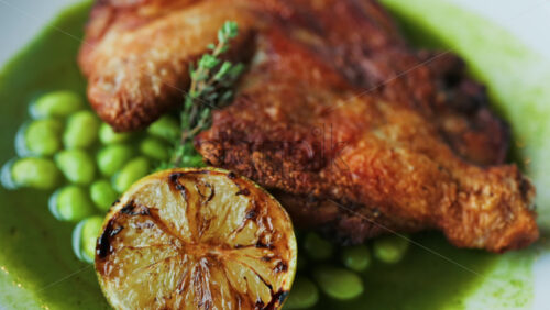 Close-up of a gourmet roasted chicken thigh served with edamame beans, a drizzle of herb-infused green sauce, and a charred lime half - Starpik Stock