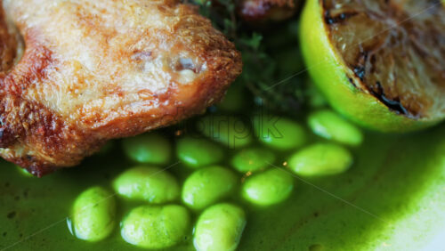 Close-up of a gourmet roasted chicken thigh served with edamame beans, a drizzle of herb-infused green sauce, and a charred lime half - Starpik Stock