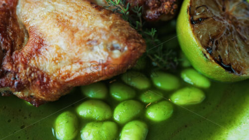 Close-up of a gourmet roasted chicken thigh served with edamame beans, a drizzle of herb-infused green sauce, and a charred lime half - Starpik Stock