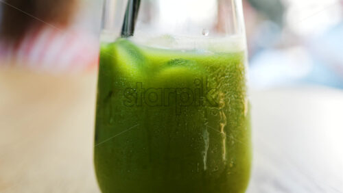 Close up of a hand mixing in a glass of an iced orange juice matcha with a black straw on a table at a cafe - Starpik Stock