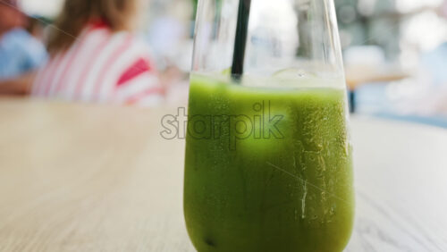 Close up of a hand mixing in a glass of an iced orange juice matcha with a black straw on a table at a cafe - Starpik Stock