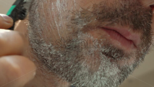 Close up of a man shaving his beard with a green and black razor - Starpik Stock