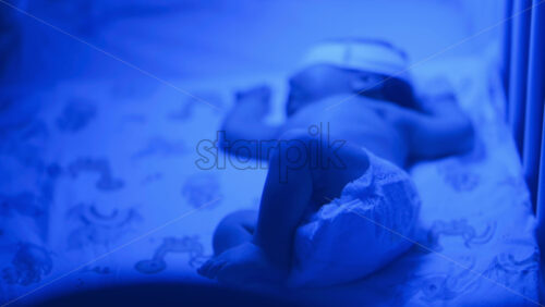 Close up of a newborn baby moving around on a crib mattress, wearing a diaper with calming blue lighting - Starpik Stock