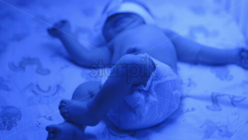 Close up of a newborn baby moving around on a crib mattress, wearing a diaper with calming blue lighting - Starpik Stock