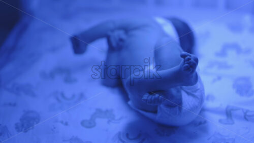 Close up of a newborn baby moving around on a crib mattress, wearing a diaper with calming blue lighting - Starpik Stock