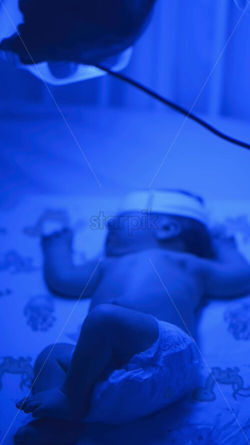 Close up of a newborn baby moving around on a crib mattress, wearing a diaper with calming blue lighting. Vertical - Starpik Stock