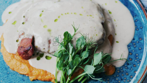 Close-up of a piece of grilled meat covered in a creamy white sauce, accompanied by a vibrant orange puree base and garnished with fresh green herbs - Starpik Stock