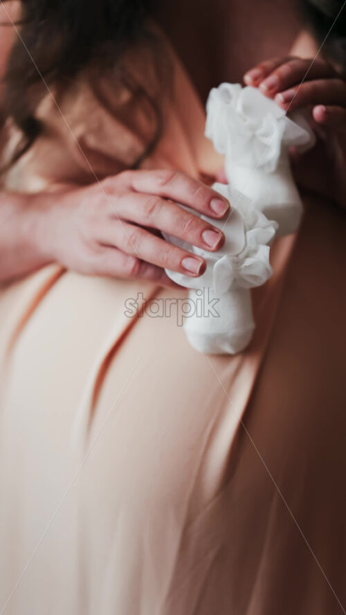 Close up of a pregnant woman holding and playing with white baby socks on her belly. Vertical - Starpik Stock