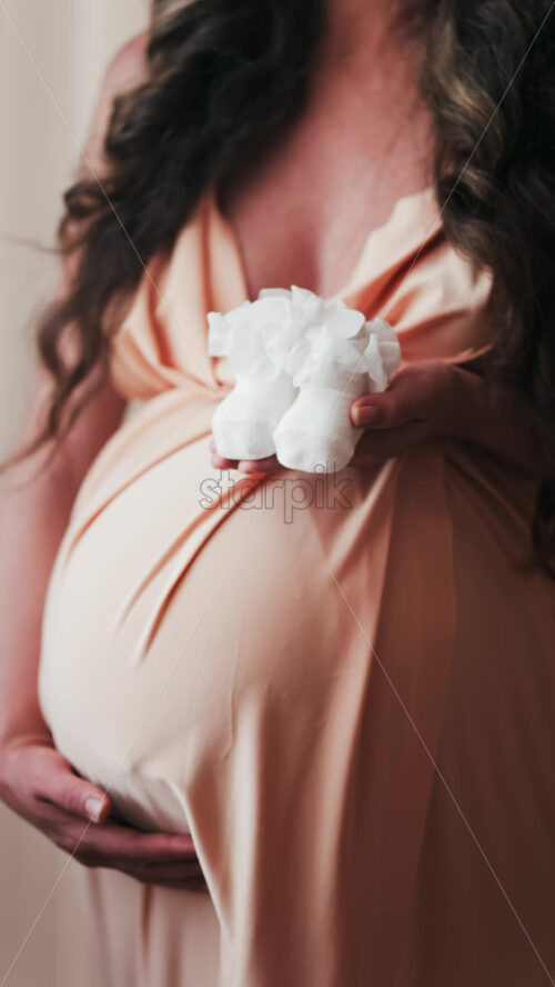 Close up of a pregnant woman holding white baby socks on her belly. Vertical - Starpik Stock