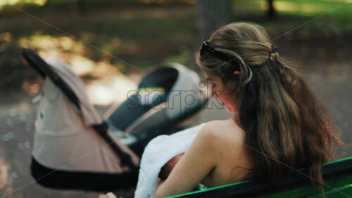 Close up of a stroller with a mother breastfeeding her baby while sitting on a park bench in the background - Starpik Stock