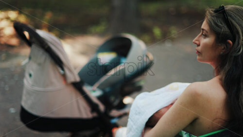 Close up of a stroller with a mother breastfeeding her baby while sitting on a park bench in the background - Starpik Stock