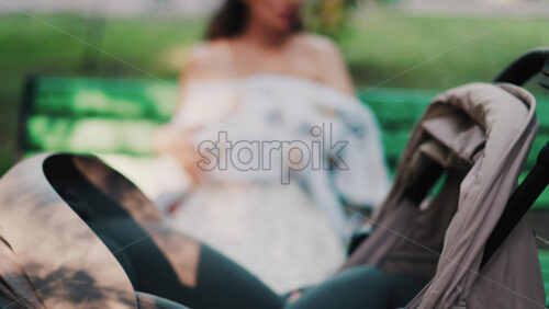 Close up of a stroller with a mother breastfeeding her baby while sitting on a park bench in the background - Starpik Stock